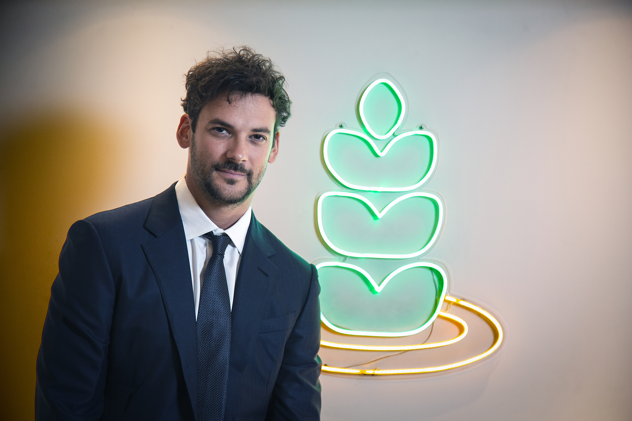 Hombre con traje formal posando frente al logotipo iluminado de una empresa agrícola con forma de espiga verde y amarilla.