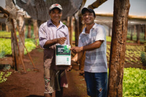 Dos agricultores sonrientes muestran un bioinsumo orgánico en una finca agroecológica con cultivos de hoja verde.