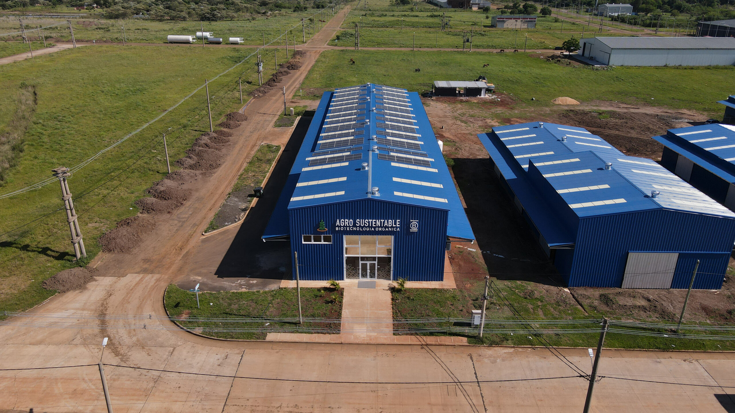 Vista aérea de la planta de Agro Sustentable en Misiones, con techo azul y paneles solares, rodeada de caminos de tierra y áreas verdes.