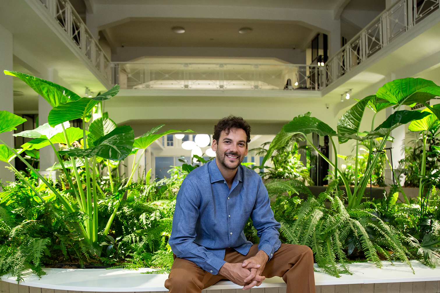 Joaquín Basanta sonríe en un entorno interior lleno de plantas tropicales, rodeado de vegetación exuberante y arquitectura moderna.