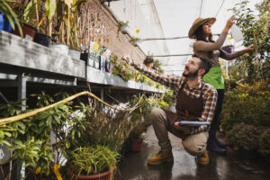 Dos personas jardineras trabajando alegremente en un invernadero, cuidando plantas en macetas y rociando agua, rodeadas de vegetación y luz natural.