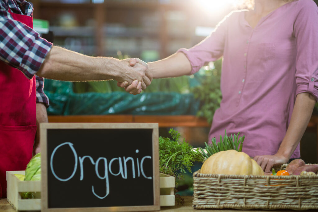 Un productor y una compradora se dan la mano frente a una canasta con vegetales frescos y un cartel que indica "Organic", en un mercado o tienda de alimentos saludables.
