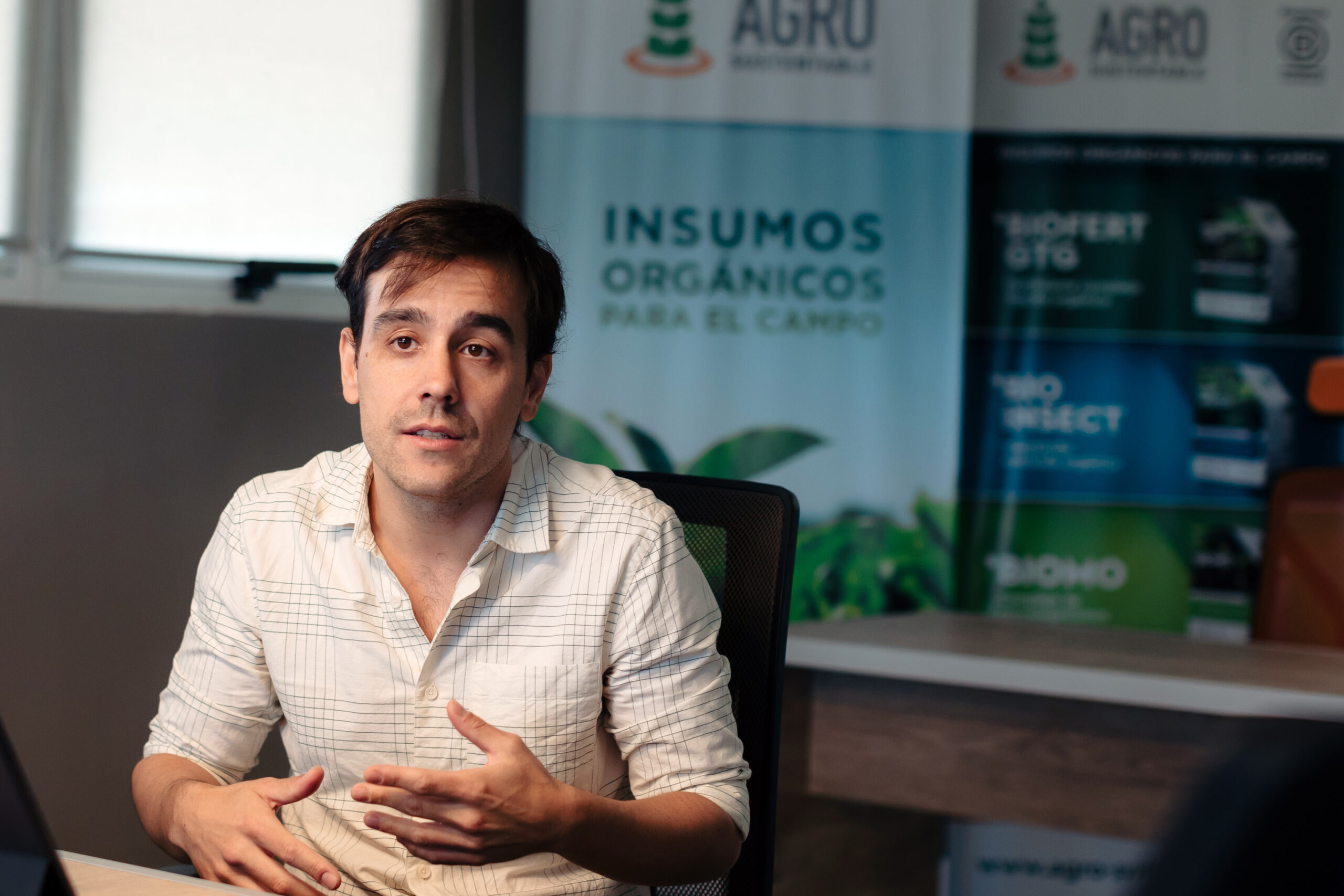 Hombre joven hablando en una oficina con fondo institucional de Agro Sustentable y carteles sobre bioinsumos.