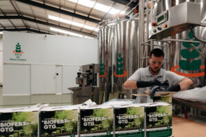 Técnico en sala de reactores de Agro Sustentable manipulando insumos para envasado de biofertilizantes BIOFERT GTG, rodeado de tanques industriales y cajas del producto.