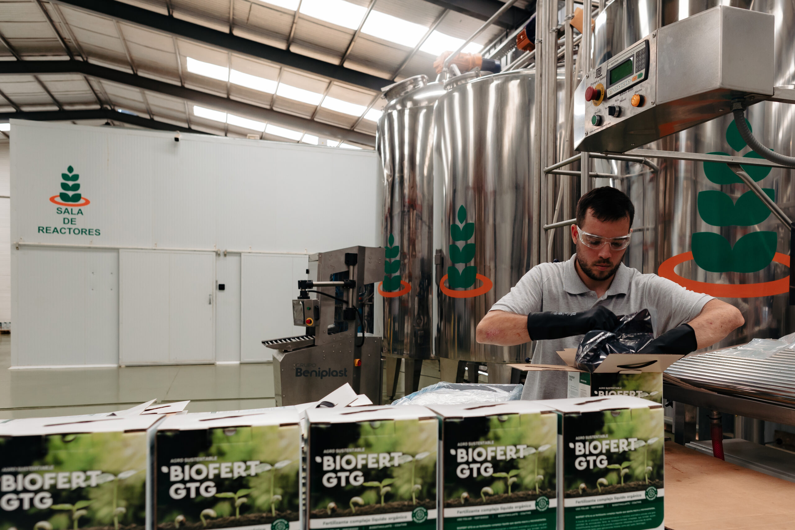 Técnico en sala de reactores de Agro Sustentable manipulando insumos para envasado de biofertilizantes BIOFERT GTG, rodeado de tanques industriales y cajas del producto.